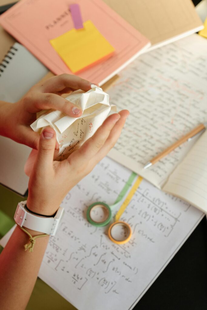 A student crumpling paper over math notes and plan books on a desk.